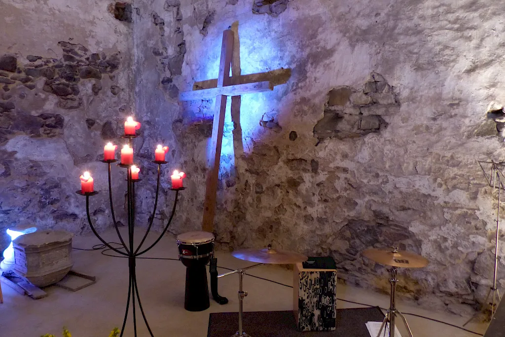 Holzkreuz vor der Bruchsteinwand im blauen Licht, davor ein Leuchter mit brennenden roten Kerzen