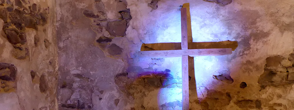 Schlichtes Holzkreuz an der Bruchsteinwand, von blauem und violettem Licht angestrahlt